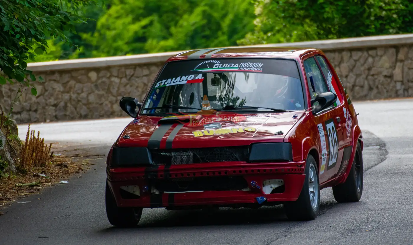 Vettura rossa numero 36 di Guida Motorsport in appoggio sull'anteriore lungo il muretto in pietra, in mezzo al verde.
