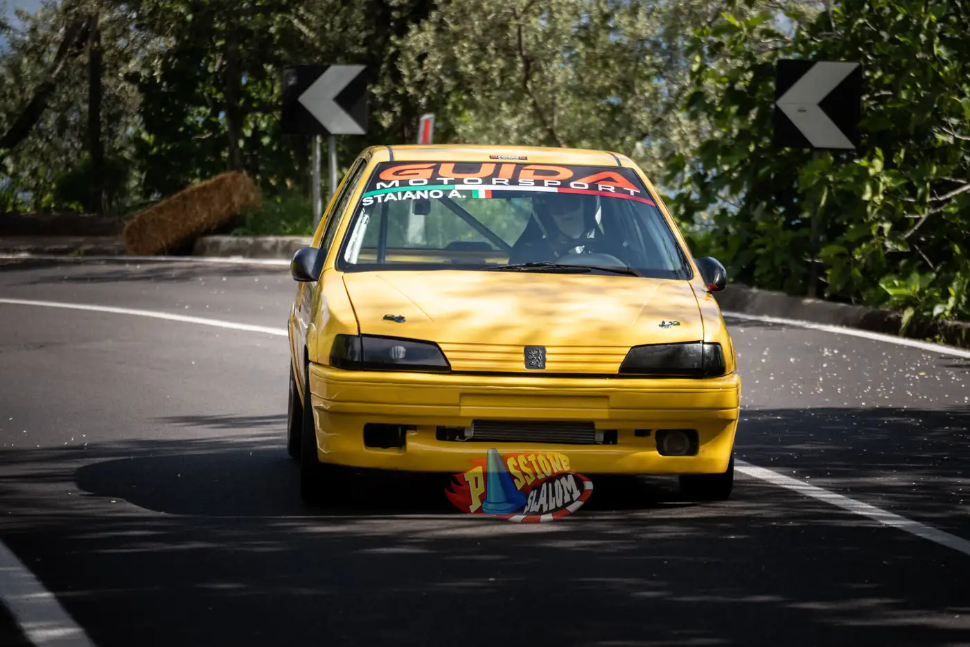 Peugeot 106 gialla di Guida Motorsport in uscita di curva, tra le frecce direzionali e la balla di fieno a bordo strada.